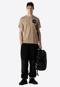 Beige bomulds T-shirt med sort logo-lomme, parret med sorte fleece og stof joggers. Sort rygsæk med justerbare stropper holdt i hånden.