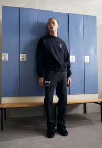 Crna dukserica s logom, uparenjena s tamnim trenirama na kojima je natpis "INTER". Glatki materijal, opušteni kroj, nosi se s crnim tenisicama u teretani.