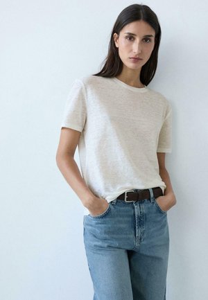 Donna con capelli lunghi e scuri che indossa una maglietta bianca a maniche corte infilata in jeans blu a vita alta, con le mani nelle tasche, in piedi contro una parete bianca.