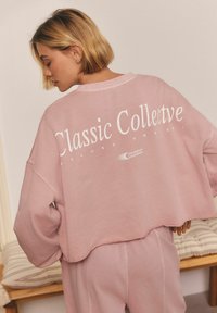 Růžová oversized mikina s kulatým výstřihem a spadlými rameny. Na zadní straně je vytištěn nápis "Classic Collective" v bílé barvě.