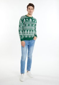 Grüner gestrickter Pullover mit weißen Wintermustern, darunter Bäume und Rehe. Kombiniert mit hellblauen Jeans und weißen Schuhen.