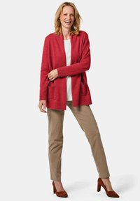 Roter Cardigan mit offener Vorderseite, gerippten Bündchen und zwei vorderen Taschen, kombiniert mit beigen Hosen und braunen Wildlederhochschuhen.