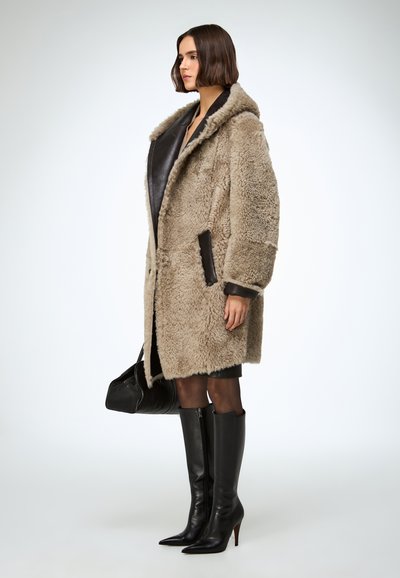 Palton din blană sintetică bej cu față din piele neagră, glugă mare, două buzunare laterale și croială lejeră, asortat cu cizme negre din piele până la genunchi.