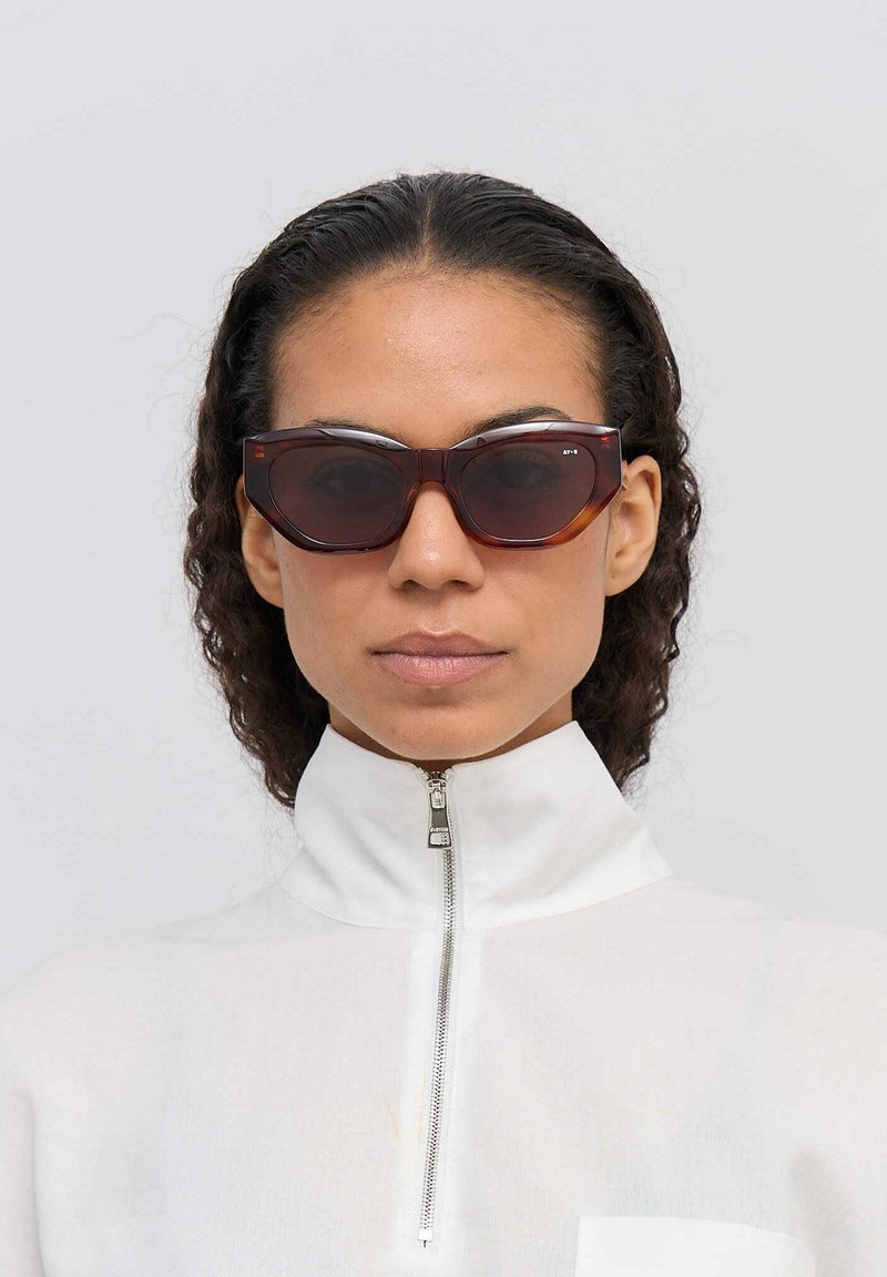 Bruna kattögon-solglasögon med en utpräglad vinkelkantad design och glansig yta. Åtföljd av en vit zip-up topp med hög krage.