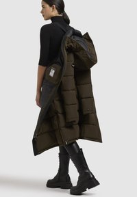 Cappotto lungo tipo puffer in verde oliva scuro con cappuccio, dotato di sezioni imbottite, chiusura con zip e numerose tasche. Dettagli in metallo nero.