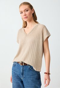 Femme portant un pull beige côtelé à col en V et manches courtes, un jean bleu avec une ceinture marron et un bracelet marron à son poignet droit.
