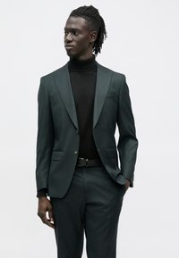 Fato verde escuro com design ajustado, casaco de um só peito e calças a combinar, combinado com camisola de gola alta preta e cinto.