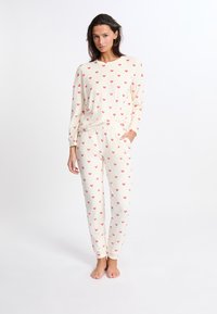 Kermanen pyjamapuku, jossa on pitkähihainen paita ja housut, jotka on koristeltu pienillä punaisilla sydänkuvioilla. Valmistettu pehmeästä kankaasta ja rentoa istuvuutta.