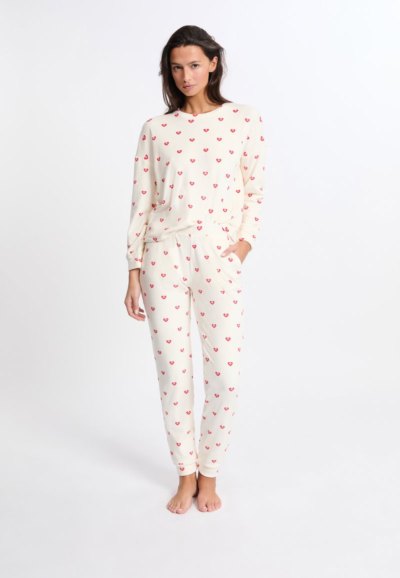 Kermanen pyjamapuku, jossa on pitkähihainen paita ja housut, jotka on koristeltu pienillä punaisilla sydänkuvioilla. Valmistettu pehmeästä kankaasta ja rentoa istuvuutta.