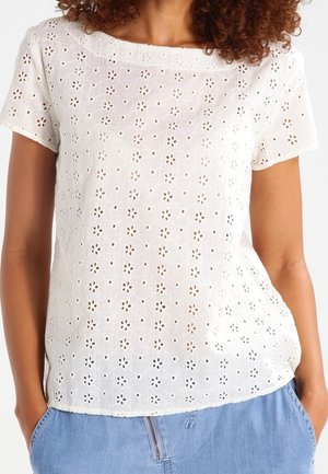 Femme portant un chemisier blanc à manches courtes avec un motif floral en découpe et un jean bleu clair, les mains dans les poches.