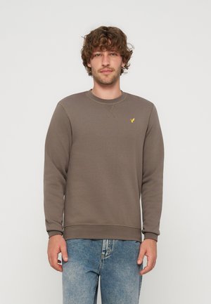 Jeune homme aux cheveux bruns bouclés portant un sweat-shirt marron avec un petit logo d'oiseau jaune et un jean bleu, debout devant un fond uni.