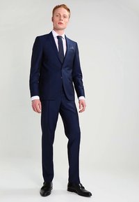 Vīrietis stāv, ģērbts tumši zilos pieguļošos uzvalkos, baltā krekla, tumšā kaklasaitē, melnās kurpēs, ar neitrālu gaiši pelēku fona.