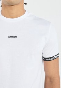 Biely bavlnený tričko s čiernym logom "URTRN" na hrudi a čierno-bielym plameňovým vzorom na rukávoch. Ľahká látka.