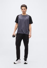 Νεαρός άνδρας που στέκεται φορώντας γκρι και μαύρο αθλητικό μπλουζάκι Puma, μαύρο παντελόνι Puma, λευκά αθλητικά παπούτσια, κρατώντας μπουκάλι νερού και ποδοσφαιρική μπάλα.