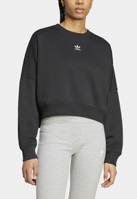 Černá zkrácená mikina s dlouhými rukávy a žebrovanými manžetami. Obsahuje bílé logo Adidas na hrudi. Spárována s světlými šedými legínami.