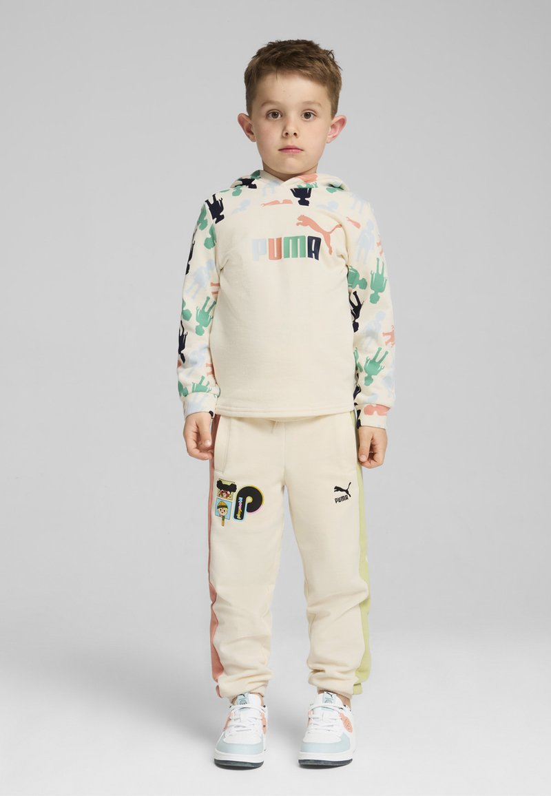 Chándal de niño que presenta una sudadera beige con estampados de animales coloridos y un logo, acompañada de pantalones de chándal beige con acentos laterales y parches.