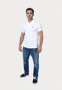 Camisa polo blanca de manga corta con cuello y un pequeño logo, combinada con jeans de denim azules y unas zapatillas náuticas navy con acentos blancos.