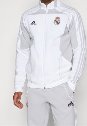 Mężczyzna ubrany w biało-szary dres Adidas z logo Realu Madryt, stojący z jedną ręką w kieszeni, a drugą opuszczoną.