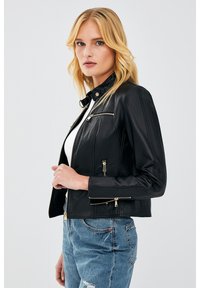 Black leather jacket with a fitted design, featuring gold hardware accents, zip pockets, and a stand collar. Worn over a white top and denim jeans.