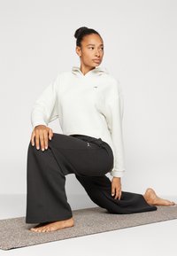 Sudadera blanca de textura con capucha, combinada con pantalones anchos negros que presentan un logotipo lateral. Ambas prendas tienen un corte relajado.