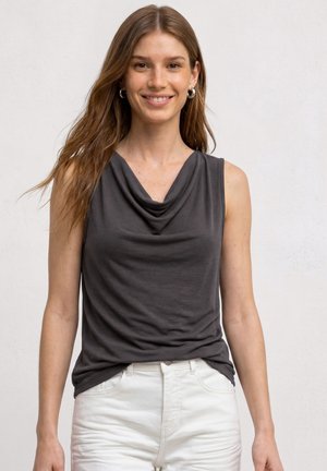 Jeune femme aux longs cheveux bruns souriante, portant un haut drapé sans manches gris foncé et un pantalon blanc sur un fond uni clair.