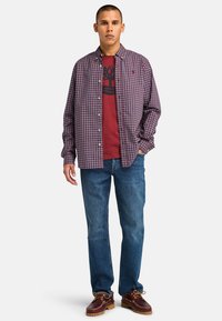 Rood en blauw geblokt overhemd over een donkerrood graphic t-shirt, gecombineerd met blauwe spijkerbroek en bruine veterschoenen. Casual uitstraling.