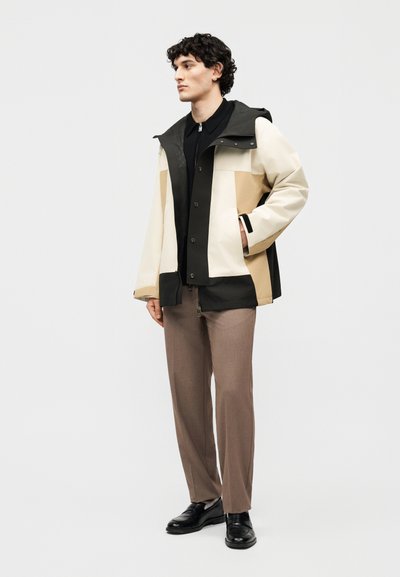 Άνδρας στέκεται με τα χέρια στις τσέπες φορώντας μπουφάν σε μπεζ, μαύρο και ταμπά χρώμα, μαύρο πουκάμισο, καφέ παντελόνι και μαύρα παπούτσια σε λευκό φόντο.