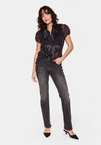 Črna prosojna bluza z nabrano sprednjo zasnovo, v kombinaciji s sivo prilegajočimi se kavbojkami in črnimi čevlji s koničastimi prsti. Gladka, rahlo teksturirana tkanina.