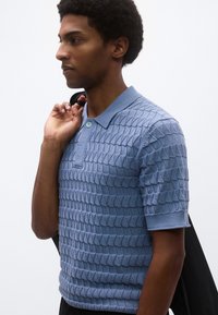 Πουλόβερ πόλο σε ανοιχτό μπλε χρώμα με υφή στο ύφασμα, κοντά μανίκια, γιακά και ένα κουμπί στον λαιμό. Φοράται με μαύρη τσάντα ώμου.