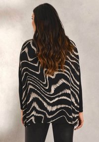 Kvinna med långt vågigt mörkt hår, iklädd en svart och beige abstrakt mönstrad långärmad topp och svarta byxor, vänd bort från kameran mot en neutral vägg.