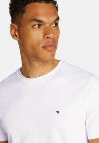 T-shirt bianco in cotone con scollo rotondo e maniche corte, con un piccolo logo rosso, bianco e blu sul petto.