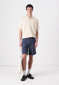 Svetlobéžový textúrovaný polo tričko, tmavomodré pruhované šortky, čierne otvorené sandále a biele ponožky. Model drží béžovú tašku.