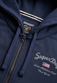 Granatowa bluza rozpinana z kapturem i sznurkami, z logo "SuperDry", flagą amerykańską oraz haftowanym napisem "State Athletics Dept New York" na przodzie.