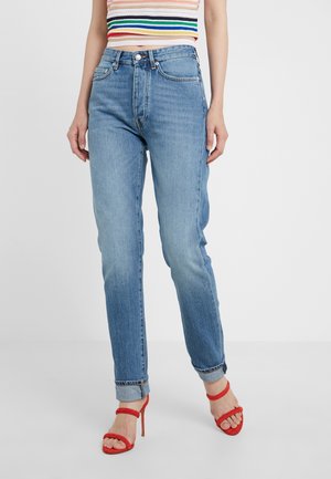 Femme portant un jean taille haute bleu avec les ourlets retroussés, un crop top sans manches à rayures colorées, et des sandales à talons hauts rouges.