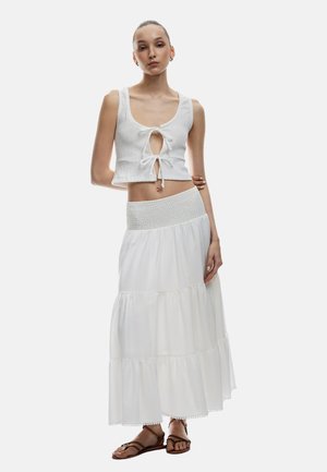 Top corto blanco con detalle de nudo en el frente y tejido texturizado, combinado con una falda maxi blanca de capas fluida. Sandalias marrones con tiras.