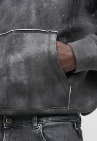 Γκρι φούτερ με υφή, με ορατές ραφές και μπροστινή τσέπη. Συνδυασμένο με σκούρες τζιν παντελόνες, που δείχνουν λεπτομέρειες από βρόχο ζώνης και κουμπί.