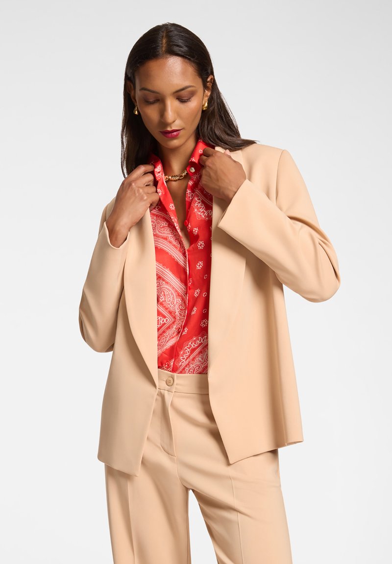 Blazer beige con design strutturato e colletti a punta, indossato sopra una camicia a motivi paisley rossi vivaci. Tessuto morbido, vestibilità ampia.