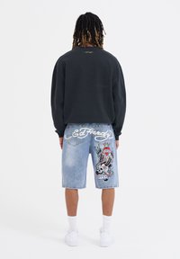 Svart sweatshirt med rundad nacke, tillsammans med ljusblå denimshorts med en färgglad tatueringsinspirerad design och broderad logotyp på baksidan.