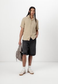 Beige kortærmet skjorte med krave, mørk denimshorts, hvide sneakers og en grå taske. Modellen står foran en hvid baggrund.