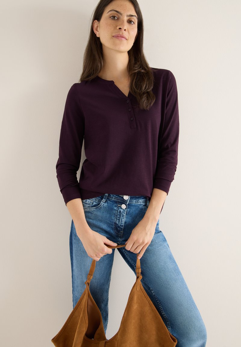 Bordeaux langærmet henley skjorte med tre-knaps placket, parret med lyseblå jeans og en lysbrun suede taske. Glat stofstruktur.