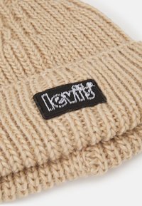 Berretto lavorato a maglia beige con una consistenza a coste spesse e un'etichetta rettangolare nera con testo ricamato bianco sul davanti.