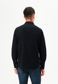 Camisa negra de manga larga hecha de una tela suave y texturizada. Presenta un yugo puntiagudo y puños abotonados, combinada con jeans azules.