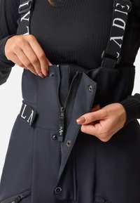 Zwarte bodysuit met een ritssluiting aan de voorkant, drukknoopsluitingen en verstelbare banden met logo-accenten. Glad, rekbaar materiaal met een getailleerd ontwerp.