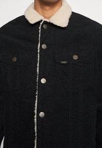 Sort kordfløjljakke med en creme-farvet faux shearling-krave. Har to brystlommer med Lee-branding og metaltryklåse.