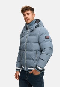Chaqueta de plumas azul claro con capucha, costuras horizontales, detalles en blanco y puños acanalados. Presenta bolsillos con cremallera y un parche de la bandera de Estados Unidos.