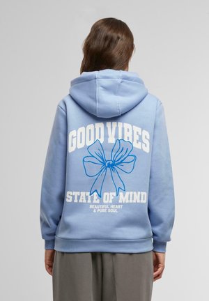 Kobieta w jasnoniebieskiej bluzie z kapturem z dużą niebieską kokardą i białym napisem „GOOD VIBES STATE OF MIND BEAUTIFUL HEART & PURE SOUL” na plecach.