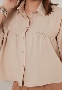 Beżowa koszula z guzikami, z kołnierzykiem, z detalem w talii oraz gładką fakturą. Projekt zawiera luźny krój i miękki materiał.