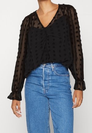 Sort bluse med gennemsigtigt, tekstureret stof, der har en V-udskæring, rynket front og puffede ærmer med elastiske mansjetter. Sammen med højtsiddende blå jeans.