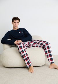 Maglia a maniche lunghe blu navy con taschino sul petto; pantaloni del pigiama a quadri in rosso, verde e bianco. Modella seduta su una sedia arrotondata e testurizzata.