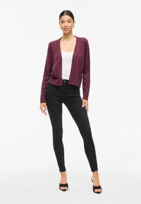 Burgundy knitted cardigan with an open front design, long sleeves, ribbed cuffs, worn over a white top and paired with black skinny jeans.
